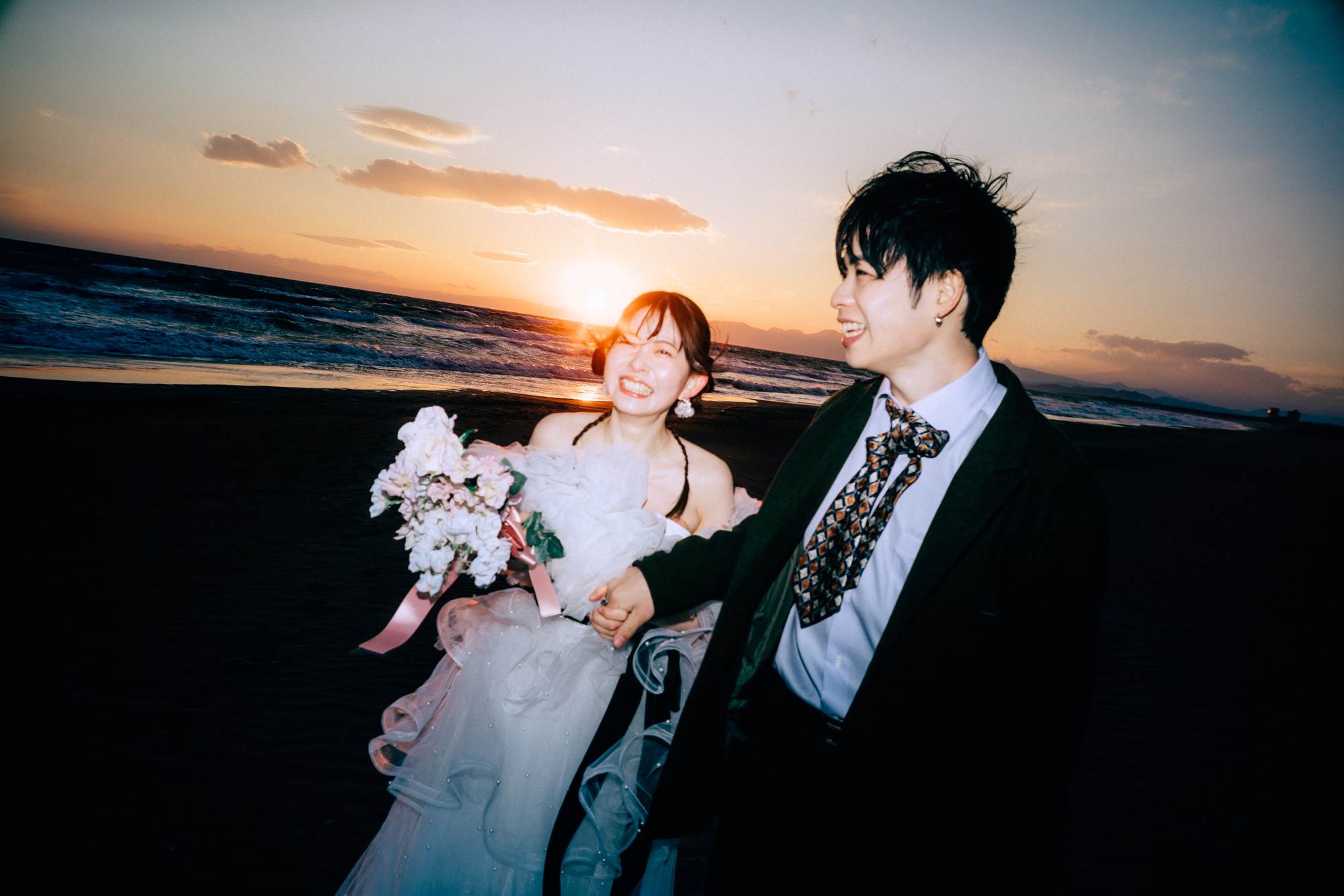七里ヶ浜の夕日をバックにブーケを持って笑う花嫁と新郎