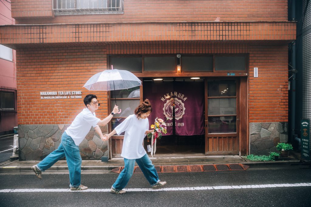 蔵前の煉瓦造りの店の前で傘をさして走るカップルの前撮り写真
